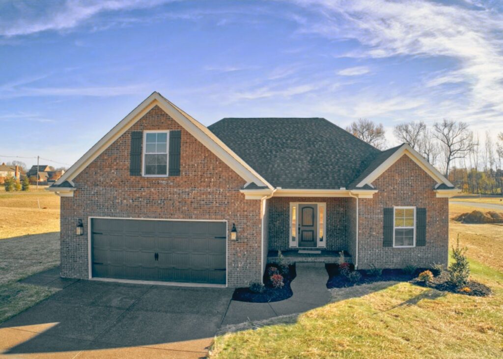 A single-story brick house by John Elpers features a dark shingle roof, double garage, and a front porch. The grassy yard is set against a clear blue sky with trees in the background.