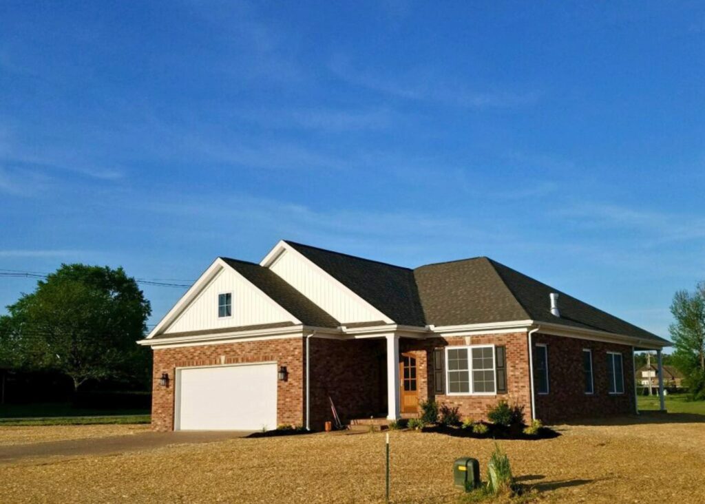 A single-story brick house by John Elpers with a white garage door, gabled roof, and wooden front door, surrounded by a gravel yard and small shrubs, under a clear blue sky.