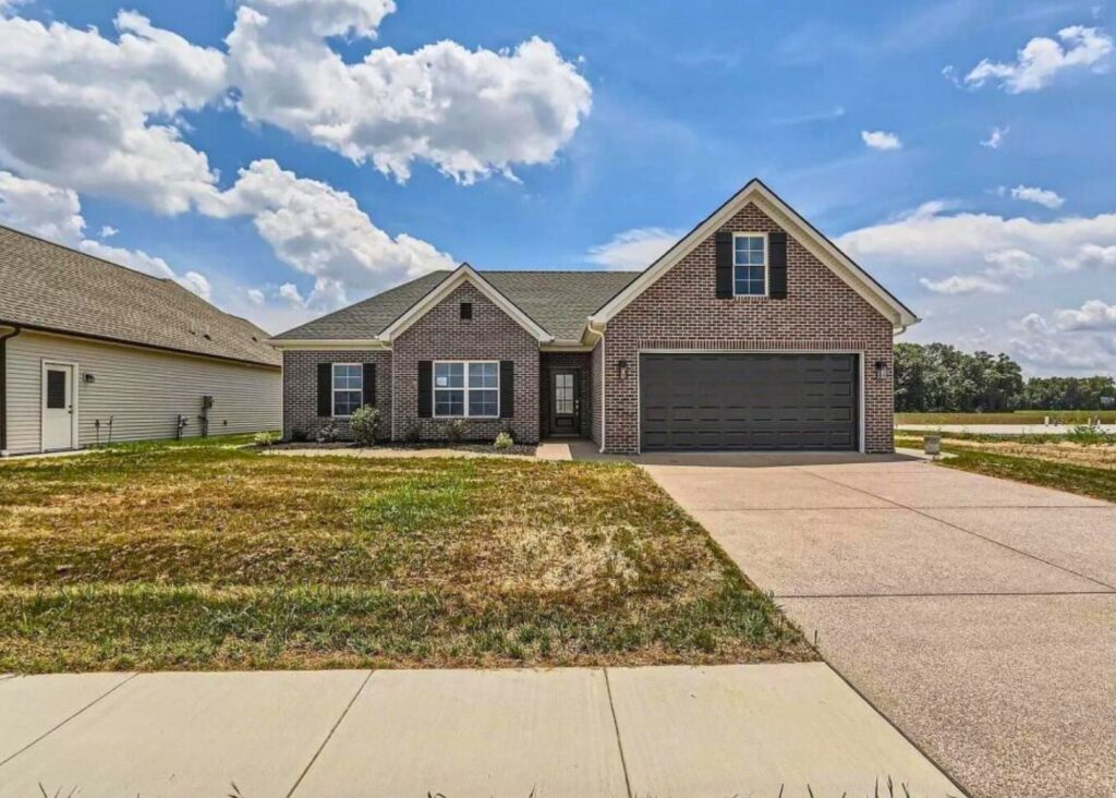 Single-story brick house by John Elpers, featuring black shutters and a dark two-car garage, set on a patchy lawn with a wide driveway. The sky is bright blue with scattered fluffy clouds.