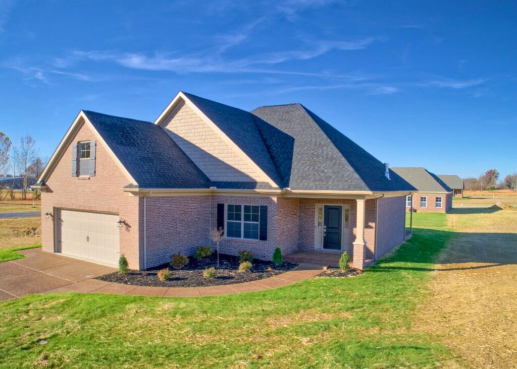 Single-story brick house by John Elpers with a dark shingle roof, attached two-car garage, small shrubs in the front yard, and a clear blue sky overhead. The home sits on a grassy lot with a sidewalk out front.