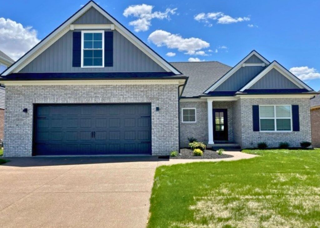 Single-story suburban house by John Elpers with gray brick exterior, black shutters, a two-car garage, gabled roof, front porch, landscaped shrubs, and a green lawn under a blue sky with scattered clouds.