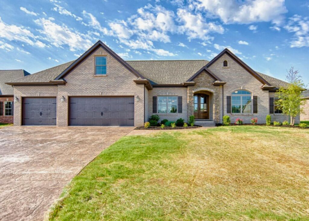 Single-story, modern brick house by John Elpers featuring a three-car garage, large front windows, manicured shrubs, and a green lawn under a partly cloudy blue sky.
