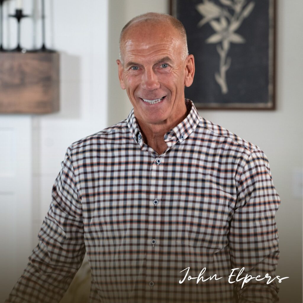 A smiling, bald man wearing a checked shirt stands indoors. In the corner, “John Elpers” is written in cursive white text. A blurred framed picture hangs behind him beside the words "Why Build New" subtly displayed on the wall.