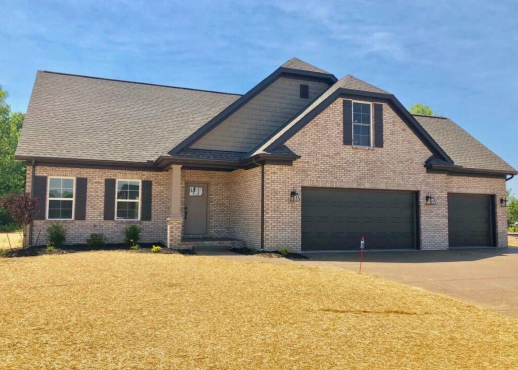 A modern single-story brick house by John Elpers with a dark roof, double garage doors, window shutters, and a covered front entry, surrounded by a freshly mulched and seeded front yard.