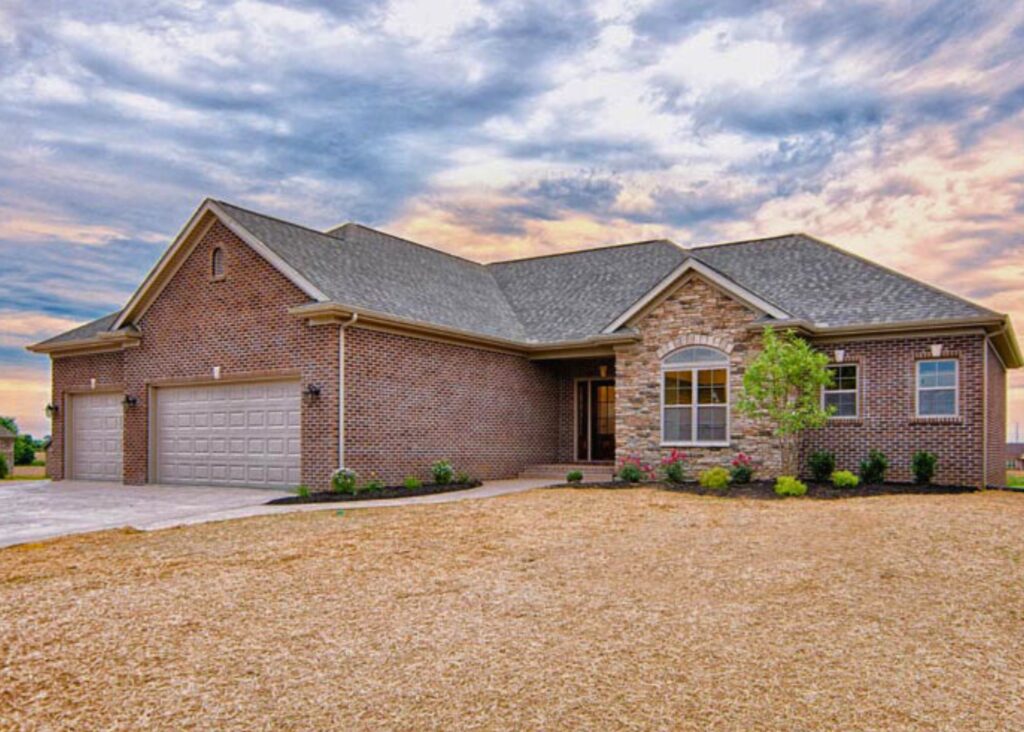A single-story brick house by John Elpers features a two-car garage, large front windows, and a small garden, all sitting under a cloudy sky at sunset with a bare front yard.