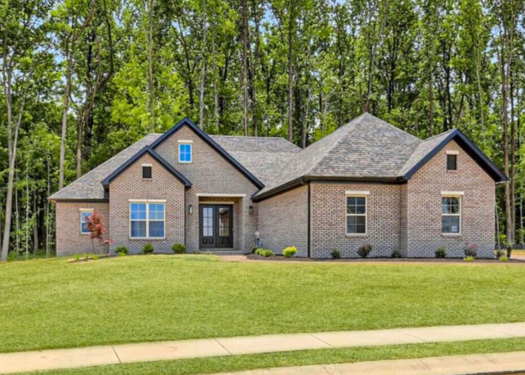 Single-story brick house by John Elpers with dark trim, arched entryway, multiple gables, and large windows, set on a neatly manicured lawn with young shrubs and trees, backed by a dense forest.