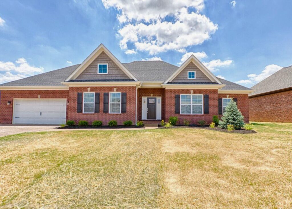A single-story brick house with a gray roof, white trim, black shutters, and a two-car garage—crafted by John Elpers. The front yard features dry grass, small bushes, a young pine tree, and a blue sky with scattered clouds.