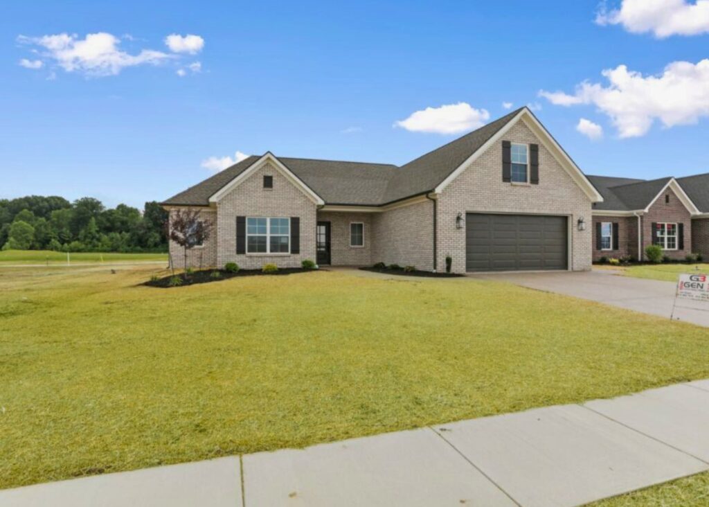 Single-story brick house by John Elpers with a two-car garage, manicured lawn, small shrubs near the foundation, and a cloudy blue sky above. A For Sale sign is visible on the front lawn near the sidewalk.