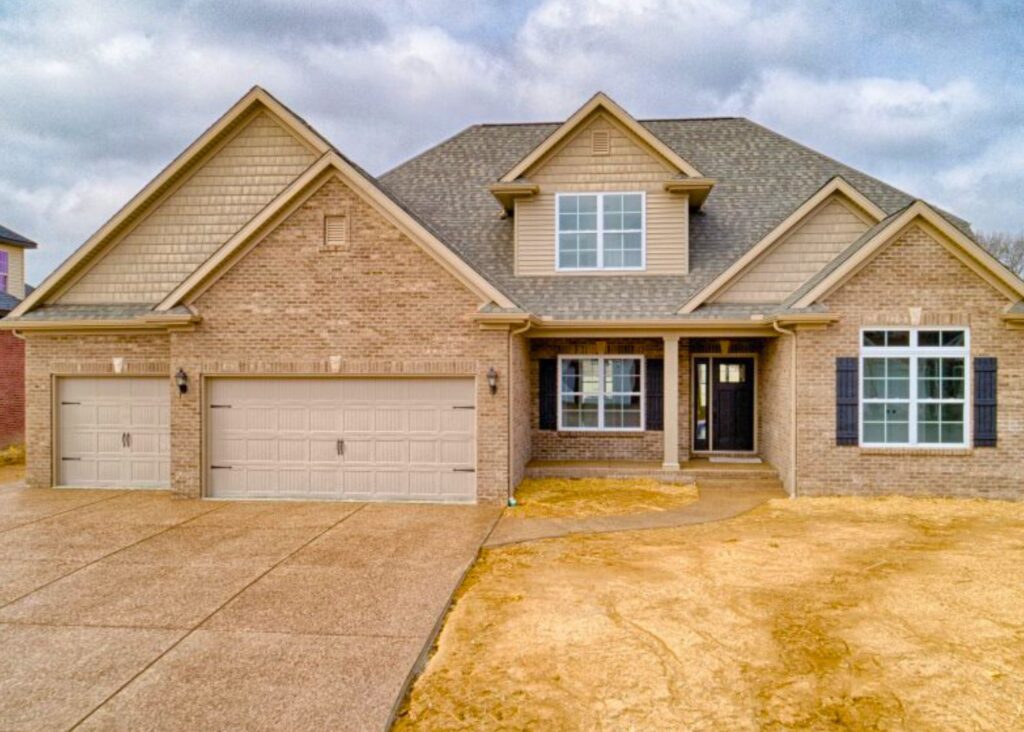 Two-story suburban house by John Elpers with light brown brick exterior, gabled roof, and attached three-car garage. Features large windows, dark shutters, double doors on a covered porch, and a bare yard awaiting landscaping.