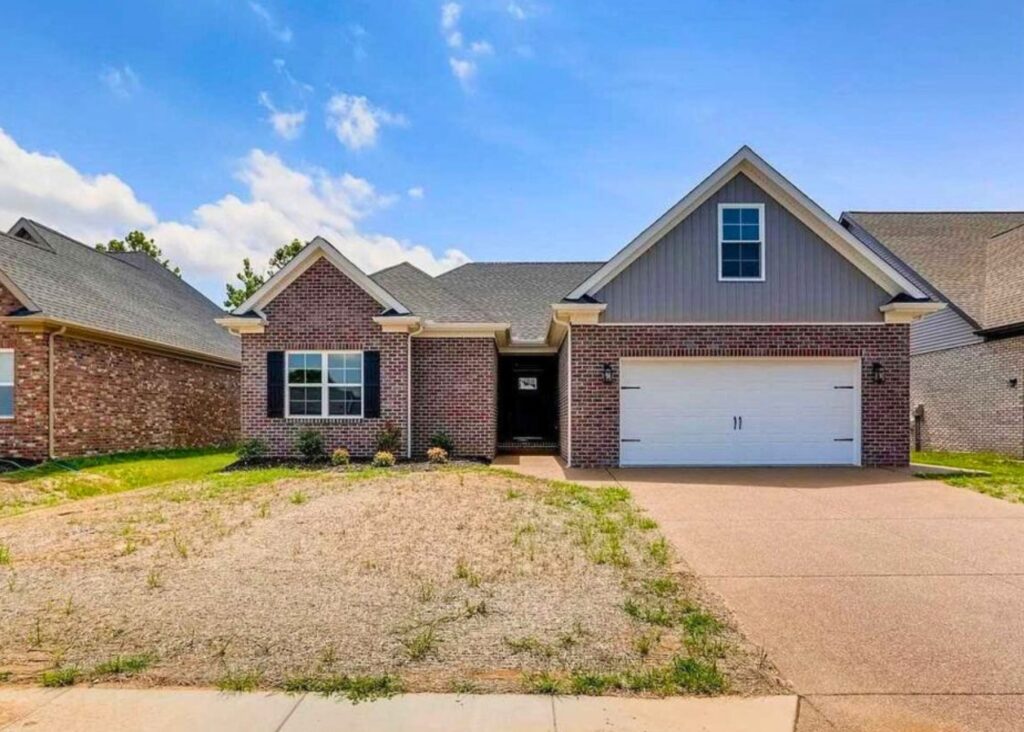 Single-story brick house by John Elpers featuring a double garage, gabled roof, and a small front yard with patchy grass. Concrete driveway and sidewalk complete the look under a mostly clear sky with a few clouds.