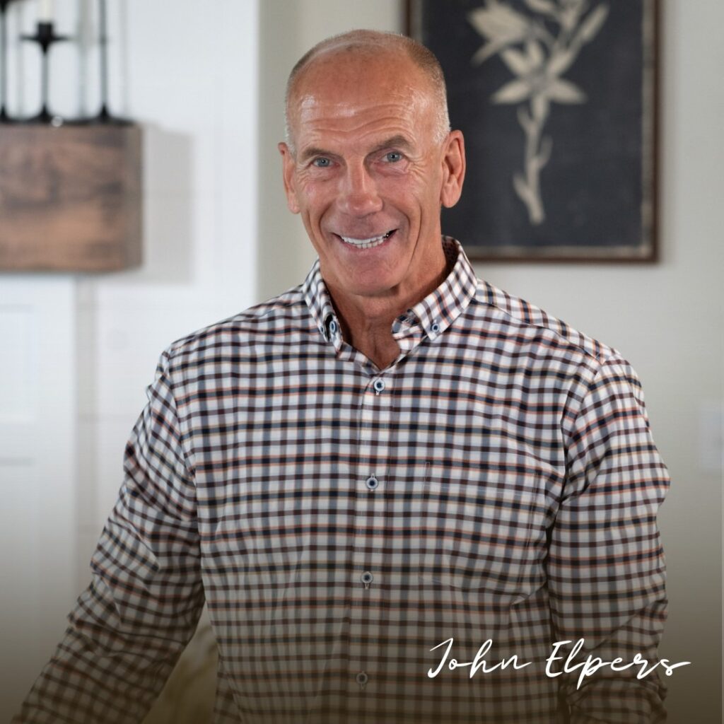 Smiling man John Elpers in a checkered button-down shirt poses indoors, with framed botanical art and warm, welcoming decor behind him.
