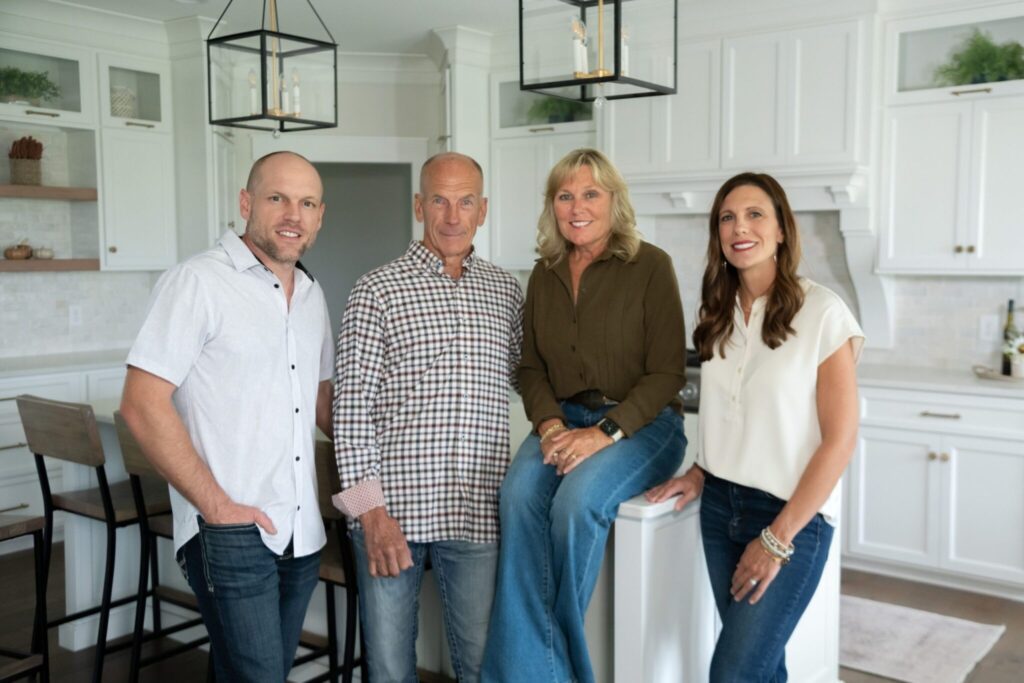 Four smiling adults pose together in a bright white kitchen beneath pendant lights, conveying warmth, friendship, and a welcoming atmosphere.