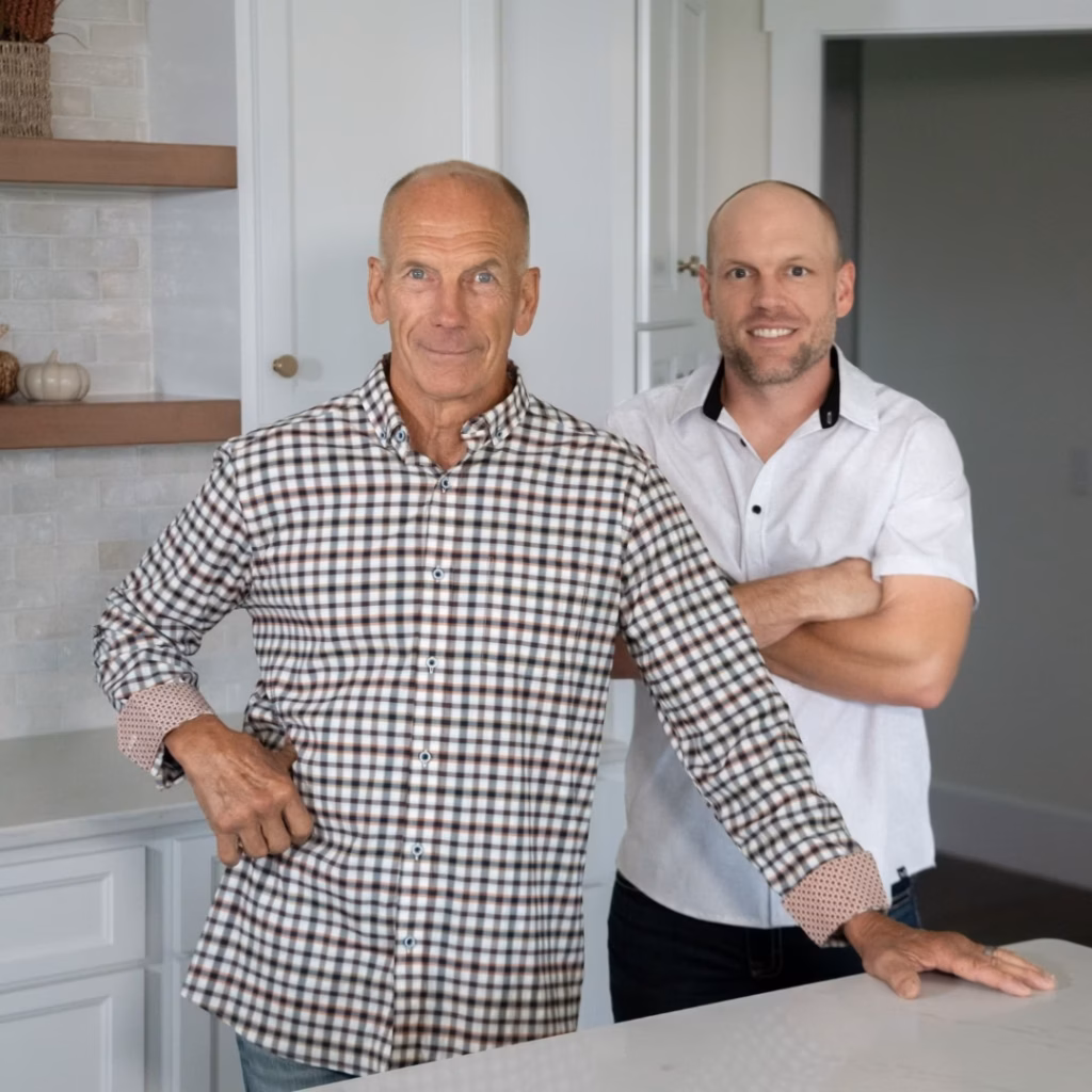 Two smiling bald men pose confidently in a bright white kitchen, one with hands on his hips and the other with crossed arms.